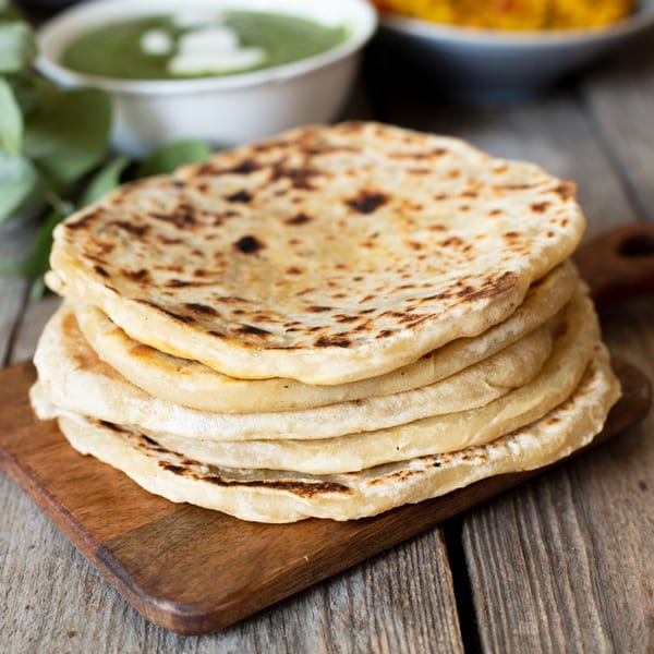 Stack of warm, grilled flatbreads on a wooden cutting board, with green sauce in background.