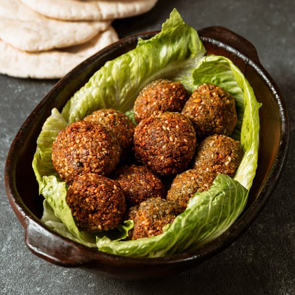A bowl of crispy falafel balls with sesame seeds, nestled on fresh lettuce leaves, with pita bread in the background.