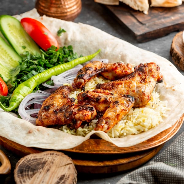 Grilled chicken wings on a bed of rice, served with fresh vegetables and flatbread.
